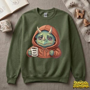 May include: Green sweatshirt featuring a cartoon dinosaur with a sad expression, wearing an orange hoodie and holding a mug that says "Nope Not Today." The dinosaur is light green with white horns. The sweatshirt is a solid, muted green color.