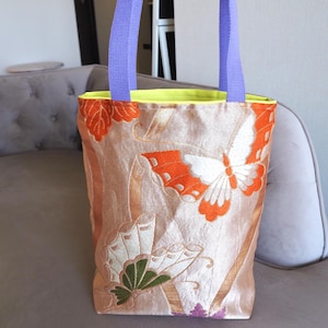 以下が含まれることがあります： オレンジ、白、緑の刺繍で蝶と葉のデザインが施されたベージュのトートバッグ。バッグの内側は黄色で、持ち手は紫です。バッグは灰色のソファの上に置かれています。