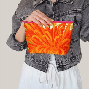 以下が含まれることがあります： 鮮やかなオレンジと赤の菊の花の刺繍が施された長方形のファスナー付きポーチ。ポーチの上部には明るいピンクのファスナーが付いており、温かみのある花柄のデザインと鮮やかなコントラストを生み出しています。