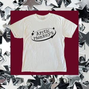 Könnte beinhalten: Ein weißes kurzärmeliges T-Shirt mit einem schwarzen Grafikdesign auf der Brust. Das Design besteht aus dem Schriftzug Arctic Monkeys in einer stilisierten Schriftart, der von einem schwarzen ovalen Rahmen mit kleinen vierzackigen Sternen umgeben ist.