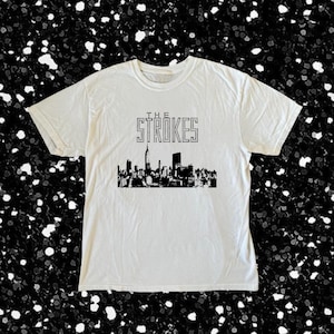 Puede incluir: Una camiseta blanca de manga corta con un estampado gráfico negro de la silueta del horizonte de Nueva York. Sobre el horizonte, el texto THE STROKES está impreso en una fuente punteada. La camiseta tiene cuello redondo y mangas cortas.
