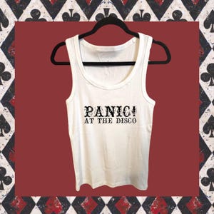 Può includere: Canotta bianca con la scritta "PANIC! AT THE DISCO" in nero. La canotta è appesa a una gruccia nera. Lo sfondo è un quadrato rosso con un bordo decorativo. La canotta ha uno scollo tondo ed è realizzata in materiale morbido.
