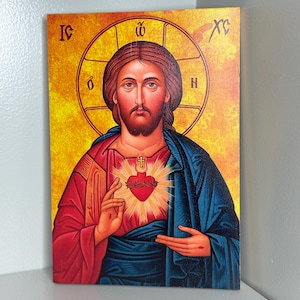 Puede incluir: Icono religioso que presenta un retrato de Jesucristo con un halo y un corazón rojo. La imagen tiene una paleta de colores cálidos con tonos rojos, azules y dorados. La obra de arte se muestra en una superficie rectangular.