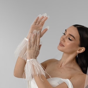 May include: A white strapless gown with a corset-style bodice, paired with sheer tulle gloves featuring white feather trim at the wrists. A matching long white tulle veil is adorned with a white ribbon and white feather trim along the bottom edge.