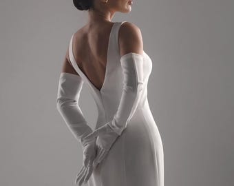 May include: A white, floor-length gown with a low back and a flowing train. The dress is paired with long, white gloves. The model is turned away from the camera, with her hands clasped behind her back.