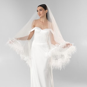 May include: A white floor-length satin gown with a strapless corset bodice and off-the-shoulder straps. A sheer white tulle veil is draped over the head and shoulders, featuring a wide trim of white feathers along the bottom edges of the veil.