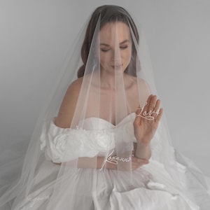 May include: A bride in a white wedding dress with a sheer veil. The veil has the word "Love" embroidered on it. The dress has off-the-shoulder sleeves and a full skirt. The bride is holding the veil in front of her.