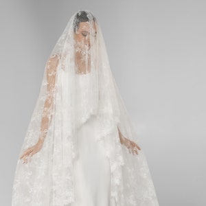 Puede incluir: Un vestido de novia blanco con una falda larga y fluida y un velo de encaje a juego. El velo está intrincadamente detallado con patrones florales y cae sobre los hombros y la espalda. El vestido tiene un diseño simple y elegante.