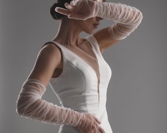 May include: A sleeveless white dress featuring a deep V-neckline with a sheer mesh insert, paired with long, ruched, semi-sheer white tulle gloves that extend past the elbows. The dress has a fitted bodice and a clean, solid white fabric construction.