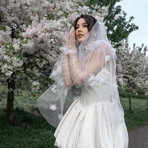 May include: A white mini dress featuring a structured corset bodice and a voluminous bubble skirt. The garment is paired with sheer, floor-length tulle gloves and a matching veil, both adorned with delicate white floral appliqu&eacute;s.