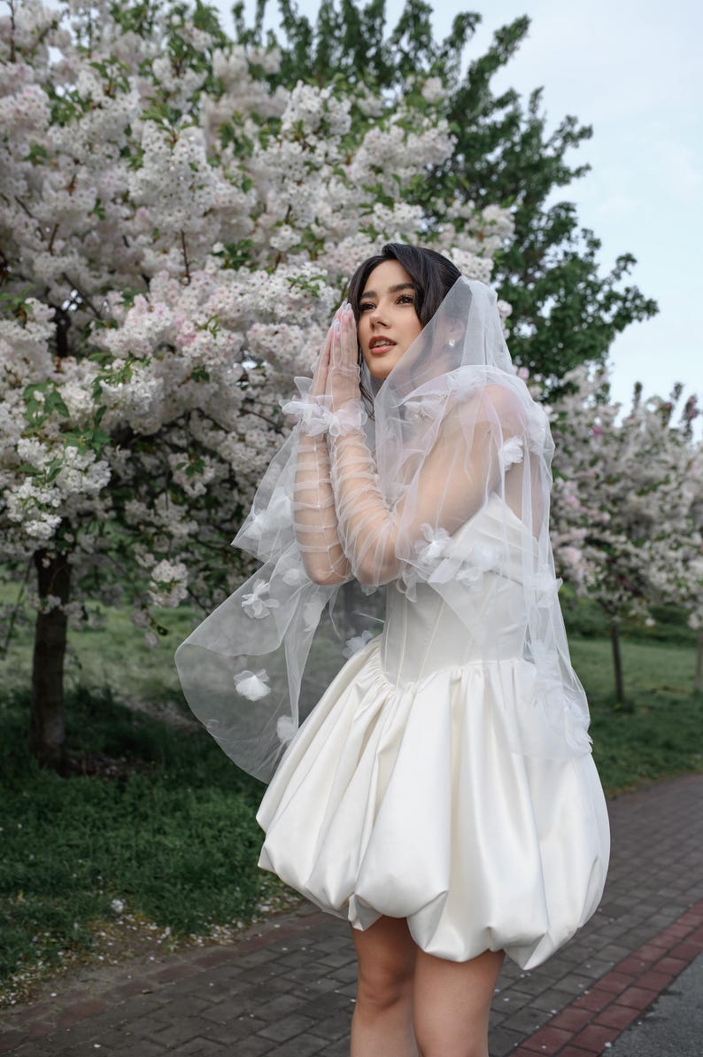 May include: A white mini dress featuring a structured corset bodice and a voluminous bubble skirt. The garment is paired with sheer, floor-length tulle gloves and a matching veil, both adorned with delicate white floral appliqu&eacute;s.