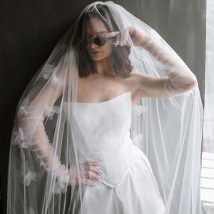 May include: A white strapless corset top and matching shorts set paired with sheer opera-length gloves and a long, flowing veil adorned with delicate white floral appliqu&eacute;s. The ensemble features clean lines and a structured bodice design.