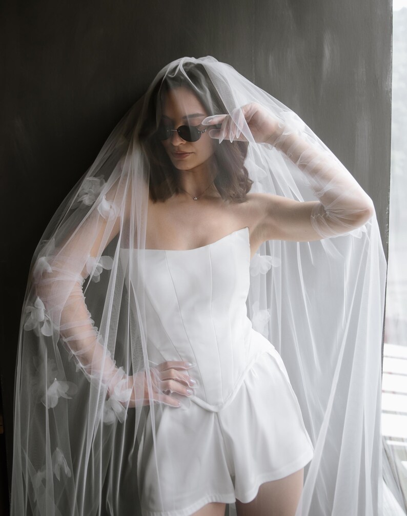 May include: A white strapless corset top and matching shorts set paired with sheer opera-length gloves and a long, flowing veil adorned with delicate white floral appliqu&eacute;s. The ensemble features clean lines and a structured bodice design.