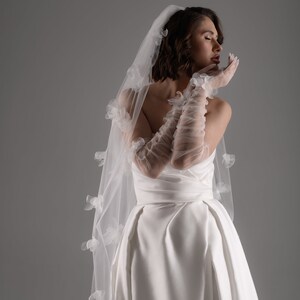 May include: A white wedding dress featuring a strapless bodice and a full skirt, paired with sheer, elbow-length ruched gloves and a long, flowing veil adorned with delicate, three-dimensional white floral appliqu&eacute;s scattered across the fabric.