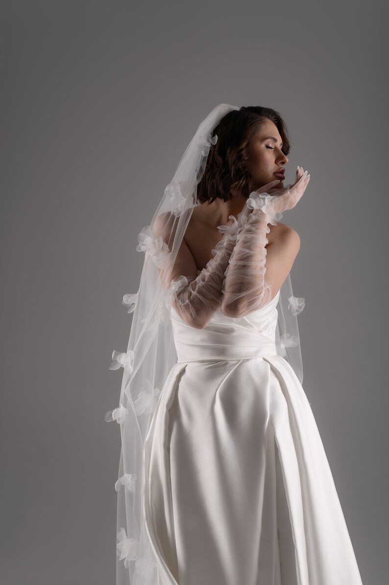 May include: A white wedding dress featuring a strapless bodice and a full skirt, paired with sheer, elbow-length ruched gloves and a long, flowing veil adorned with delicate, three-dimensional white floral appliqu&eacute;s scattered across the fabric.