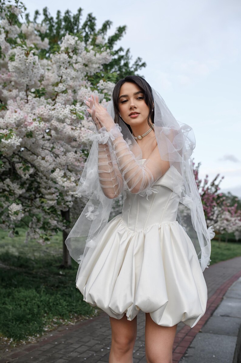 May include: A white strapless mini dress with a structured corset bodice and a voluminous bubble skirt. The ensemble includes sheer, ruffled elbow-length gloves and a matching veil adorned with small white floral appliqu&eacute;s, paired with a pearl necklace.