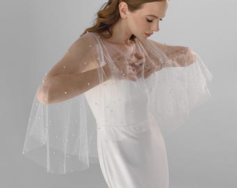 Puede incluir: Una capa corta de tul blanco transparente con perlas blancas dispersas por toda la tela. La capa tiene una silueta amplia y fluida que cae sobre los hombros y la parte superior de los brazos, sobre un vestido blanco sencillo, sin mangas y largo.