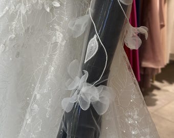 May include: A white wedding dress with intricate lace detailing and floral embellishments. The dress features a sheer sleeve adorned with delicate white leaf and flower accents. The mannequin's arm is visible, showcasing the sleeve's design.