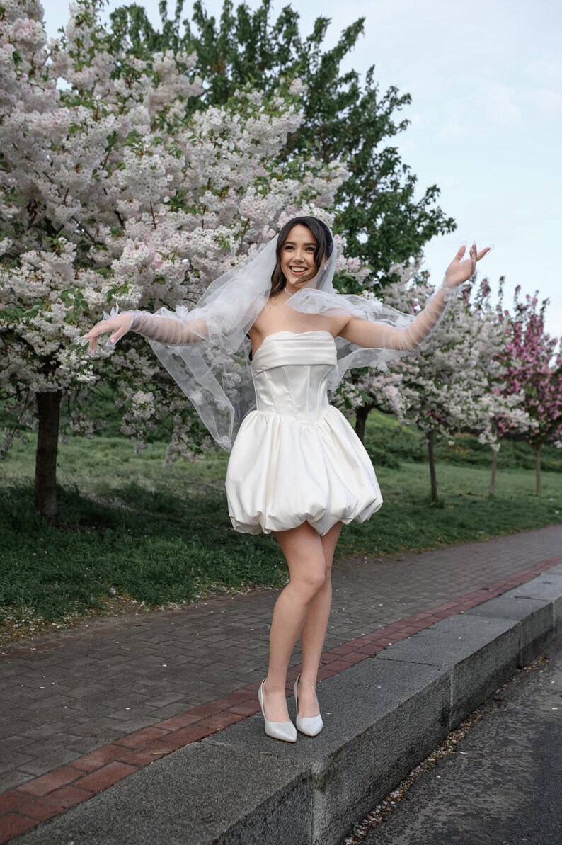 May include: A strapless white mini dress featuring a structured bodice and a voluminous bubble skirt. The outfit includes a sheer white veil with floral appliqu&eacute;s and matching sheer arm-length gloves. The ensemble is completed with white pointed-toe heels.