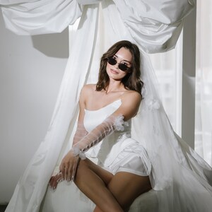 May include: A white strapless corset top paired with matching white shorts, sheer tulle opera-length gloves with floral appliqu&eacute;s, a long veil with floral details, and dark oval sunglasses. The ensemble is styled with light-coloured ankle-strap heels.