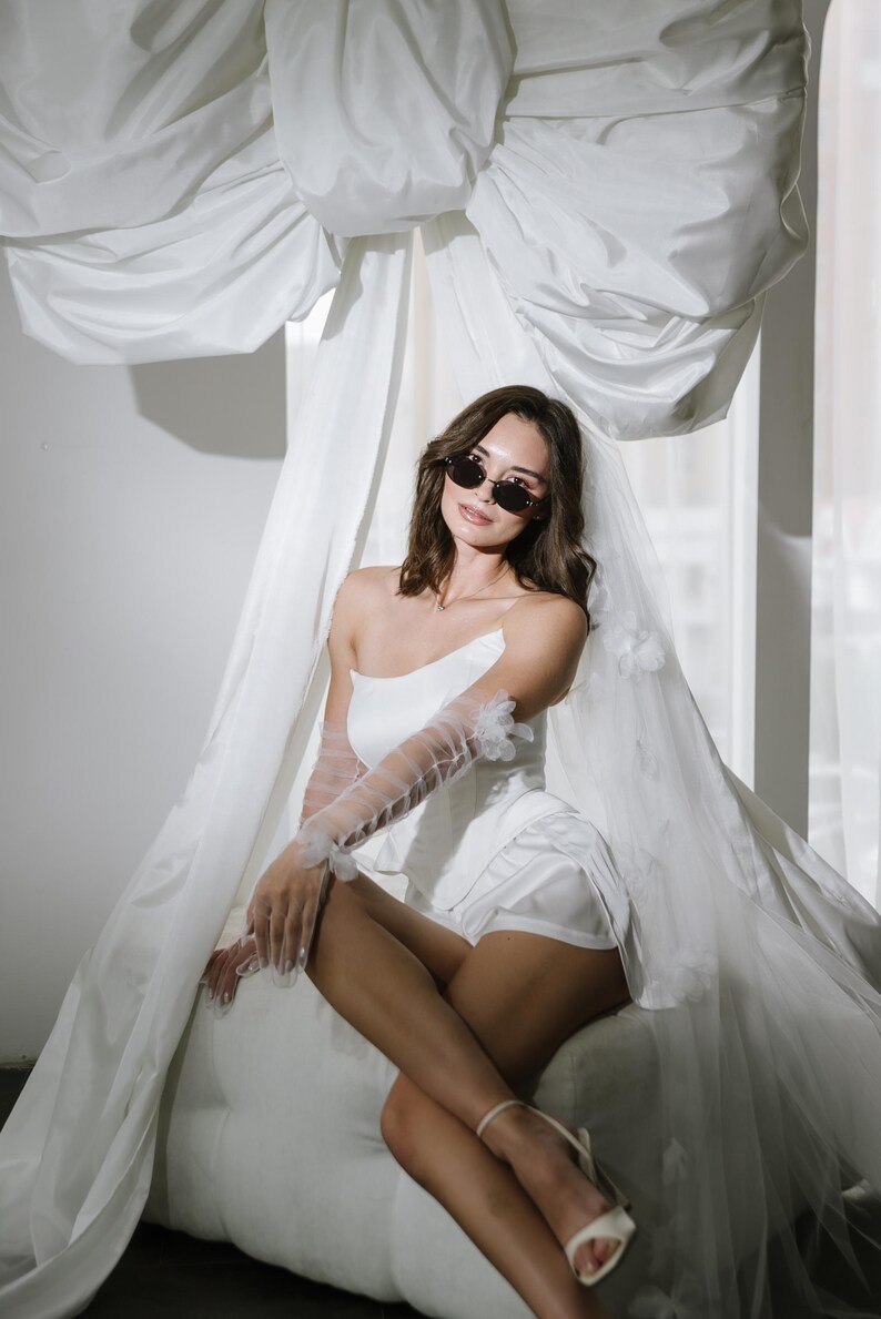 May include: A white strapless corset top paired with matching white shorts, sheer tulle opera-length gloves with floral appliqu&eacute;s, a long veil with floral details, and dark oval sunglasses. The ensemble is styled with light-coloured ankle-strap heels.