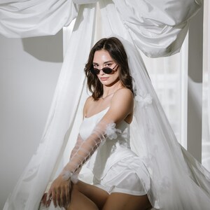 May include: A white satin corset top and matching shorts set paired with sheer, elbow-length tulle gloves featuring small floral appliqu&eacute;s. A long, flowing white veil with matching floral details is draped over the head and shoulders.