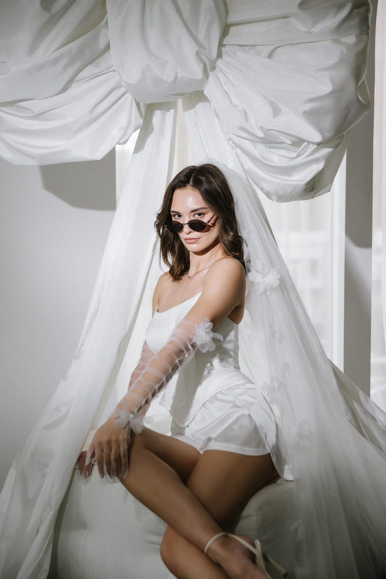 May include: A white satin corset top and matching shorts set paired with sheer, elbow-length tulle gloves featuring small floral appliqu&eacute;s. A long, flowing white veil with matching floral details is draped over the head and shoulders.