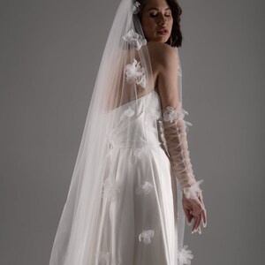 May include: A white strapless gown paired with a long, sheer veil adorned with scattered white fabric floral appliqu&eacute;s. The ensemble includes matching sheer, lace-up arm sleeves featuring similar floral details along the length of the arms.
