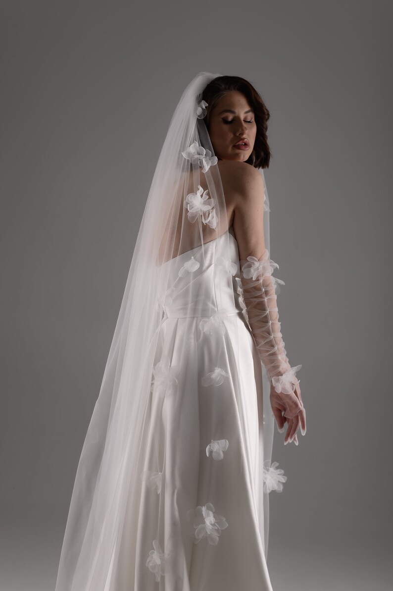 May include: A white strapless gown paired with a long, sheer veil adorned with scattered white fabric floral appliqu&eacute;s. The ensemble includes matching sheer, lace-up arm sleeves featuring similar floral details along the length of the arms.
