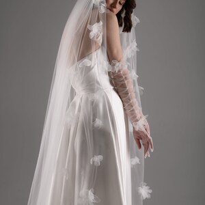 May include: A white wedding dress with a smooth satin bodice and a full skirt, paired with a long, sheer tulle veil adorned with scattered white fabric flower appliqu&eacute;s and matching sheer, lace-up opera-length gloves with floral details.