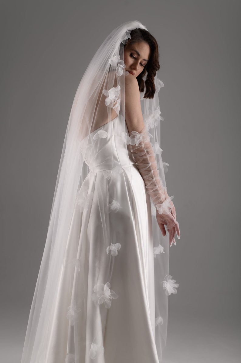 May include: A white wedding dress with a smooth satin bodice and a full skirt, paired with a long, sheer tulle veil adorned with scattered white fabric flower appliqu&eacute;s and matching sheer, lace-up opera-length gloves with floral details.
