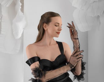 May include: Sheer black opera-length gloves featuring delicate fabric flower embellishments at the wrists. The gloves are constructed from a translucent, fine mesh material that extends past the elbows, paired with a black satin bodice dress.