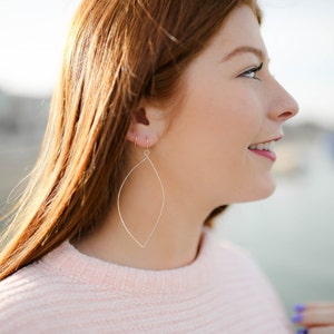 May include: A woman wearing a pink sweater with gold leaf-shaped earrings. The earrings are long and dangle from her earlobes.