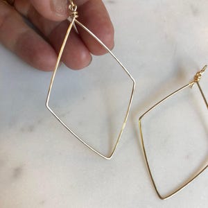 May include: A pair of gold-toned wire earrings shaped like elongated diamonds. Each earring features a thin, continuous metal wire bent into a geometric rhombus form, secured at the top with a small, tightly coiled wire loop for attachment.