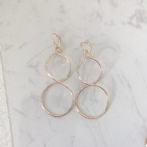 May include: A pair of gold-toned wire earrings featuring a double-loop design. Each earring consists of two interconnected circular wire hoops suspended from a simple hook-style ear wire, creating a continuous figure-eight silhouette.
