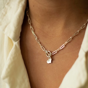 May include: Silver chain necklace with a small heart pendant. The necklace features a toggle clasp and a delicate, elongated link design. The heart pendant is smooth and polished, adding a touch of romance to the jewelry.