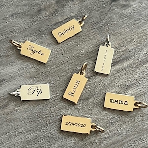 May include: Seven rectangular gold-toned metal charms with jump rings, each engraved with unique text including the names Quincy, Tagaloa, Pip, Roux, and mama, the word simplify, and the date 2/24/2020, arranged in a scattered formation.