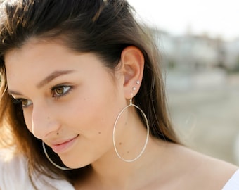 May include: A pair of large, thin, circular silver hoop earrings. The earrings feature a simple, minimalist wire design that hangs from the earlobe. The silver metal has a smooth, polished finish that reflects light, creating a bright appearance.