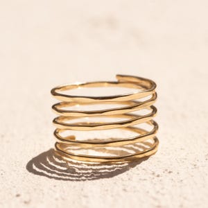 May include: A gold-toned ring featuring a multi-coil design with five distinct, stacked circular bands. The metal surface has a subtle hammered texture, creating a reflective finish that catches the light across its continuous, spiraling structure.