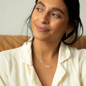 May include: A woman wearing a white shirt with a silver chain necklace with three small pearl beads.