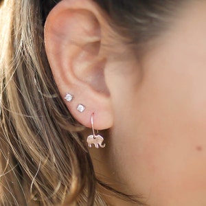 May include: A close-up of a person's ear with three earrings. The bottom earring is a silver dangle earring with a small elephant charm. The other two earrings are small silver studs.