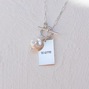 May include: A silver chain necklace featuring a toggle clasp, a single white baroque pearl, and a rectangular silver pendant engraved with the word warm in a black serif font. The necklace is composed of elongated oval links in a metallic finish.