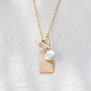 May include: A gold-toned necklace featuring a delicate chain with elongated links. The pendant consists of a circular toggle clasp, a smooth rectangular gold-toned tag, and a single white, irregularly shaped freshwater pearl charm.