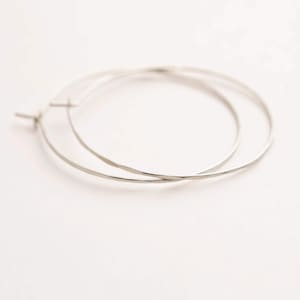 May include: A pair of thin, circular silver hoop earrings with a smooth, polished finish. The earrings feature a simple wire construction with a standard post and latch closure mechanism, designed as a continuous, lightweight metallic loop.