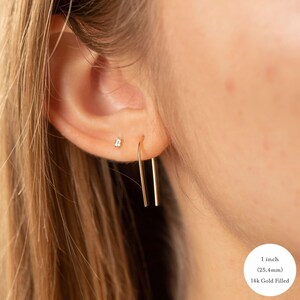 May include: A 14k gold filled U-shaped earring hanging from an earlobe, featuring a smooth, minimalist wire design. The earring measures 1 inch in length. A small, clear stud earring is also visible in the upper part of the earlobe.