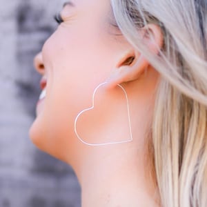 May include: A large, thin, silver-toned heart-shaped hoop earring made of a slender wire. The earring features a simple, open-frame design that forms a clear heart outline, suspended from the earlobe with a minimalist, lightweight construction.