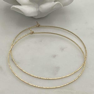 May include: A pair of thin, circular gold hoop earrings with a textured, shimmering surface. Each earring features a simple wire closure mechanism at the top, designed as a continuous loop that maintains a delicate and lightweight appearance.