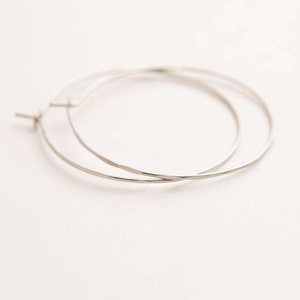 May include: Two silver-colored hoop earrings on a white background. The earrings are thin and circular, with a hammered texture. The hoops are slightly overlapping, and the earring posts are visible.