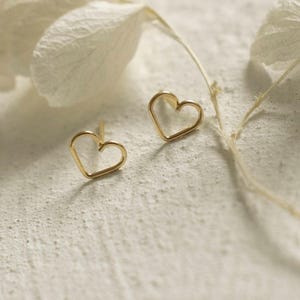May include: A pair of gold-toned stud earrings, each shaped as a simple, open-wire heart outline. The earrings feature a delicate, thin metal construction with a polished finish, designed as minimalist heart-shaped studs for ear piercings.