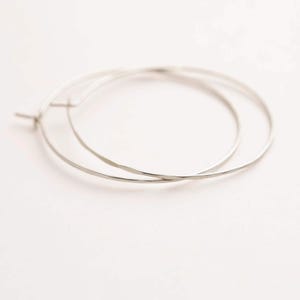 May include: A pair of thin, circular silver hoop earrings with a smooth, polished finish. The earrings feature a simple wire construction with a small, integrated post and hinge closure mechanism, creating a clean and continuous circular silhouette.